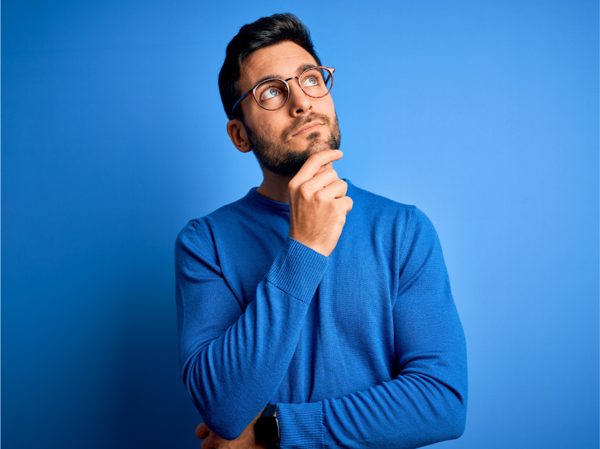 Male wearing glasses in blue shirt, in front of blue background, with hand on chin looking up and thinking